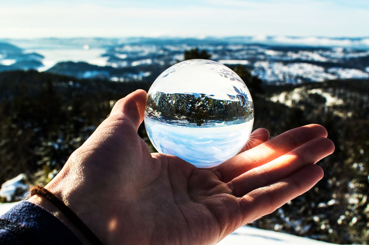 A close-up of a hand holding a crystal ball outdoors, reflecting a scenic landscape.
